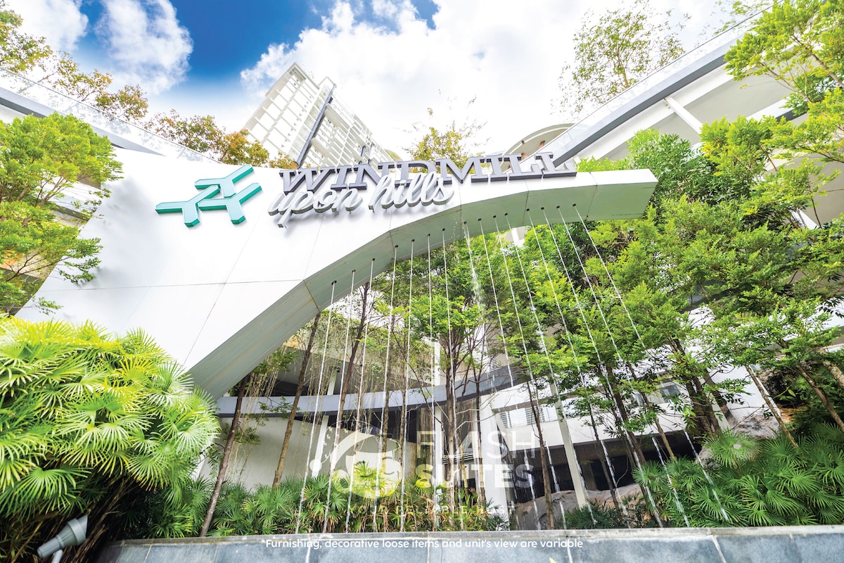 The entrance of a modern building is framed by lush greenery, with a prominent sign displaying 'Wind River Upon Hills.' Water features cascade down in front, creating a serene atmosphere. The structure rises against a backdrop of trees and a clear sky.