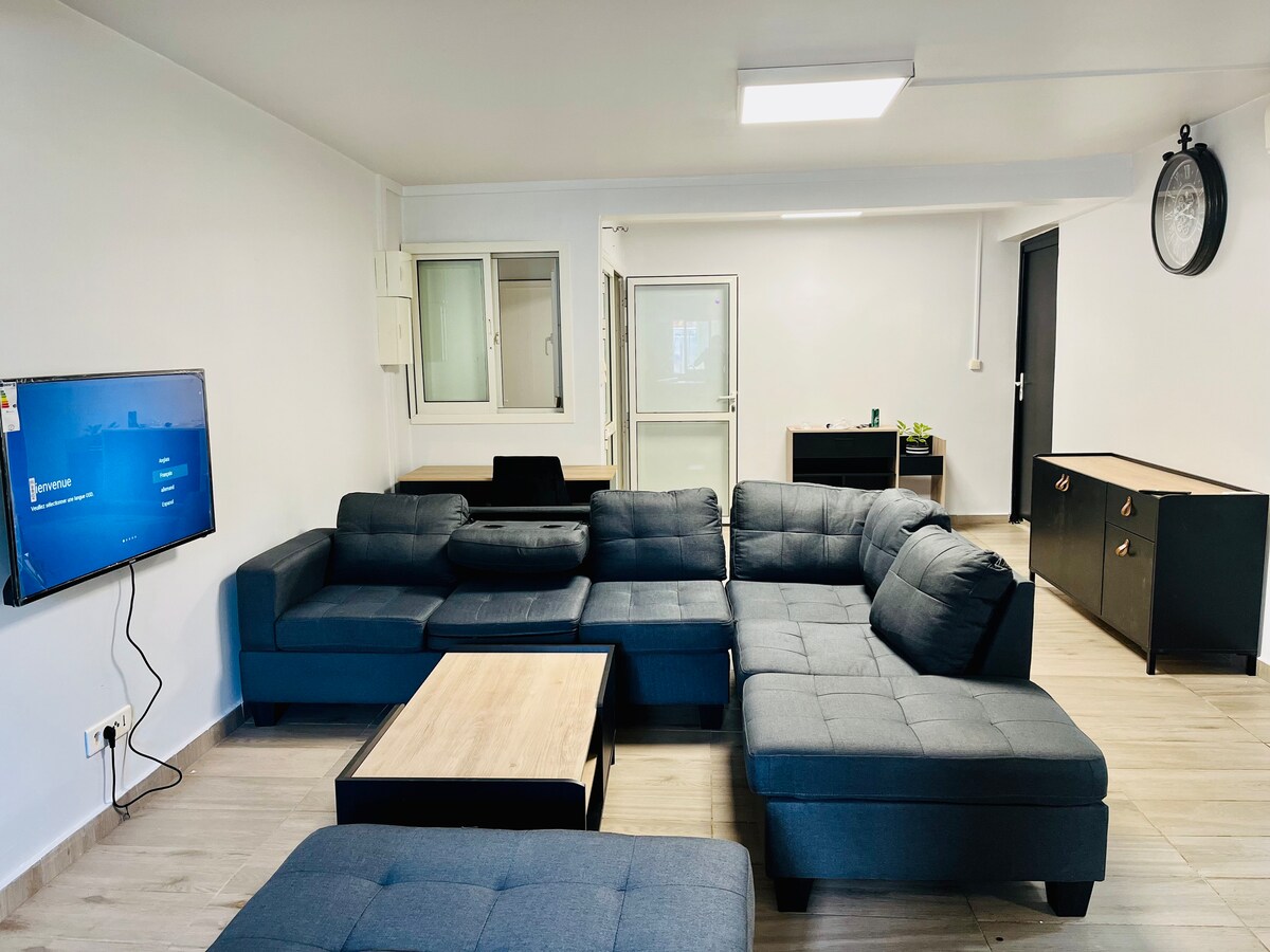 A spacious living area features a large, dark sectional sofa with a central coffee table. A flat-screen television is mounted on the wall, and ample natural light enters through large windows. A modern desk and cabinet are also present in the background.