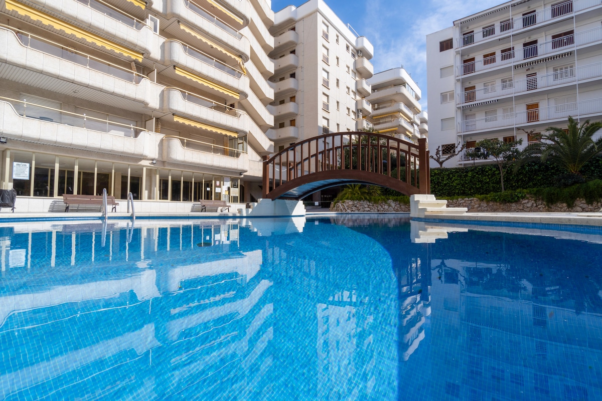A community pool is visible with clear blue water, reflecting the surrounding buildings. A wooden bridge crosses over a section of the pool, while palm trees and landscaped greenery add a natural touch to the environment.