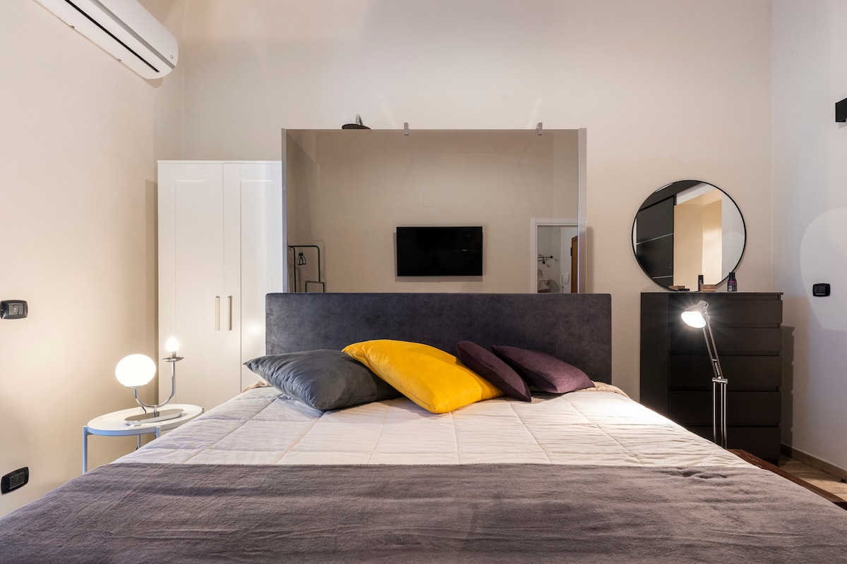 A spacious bed is centered in the room, adorned with colorful pillows in yellow, purple, and dark gray. A modern mirror and a sleek TV are visible on the wall, while stylish lighting fixtures enhance the design. Air conditioning is also present.