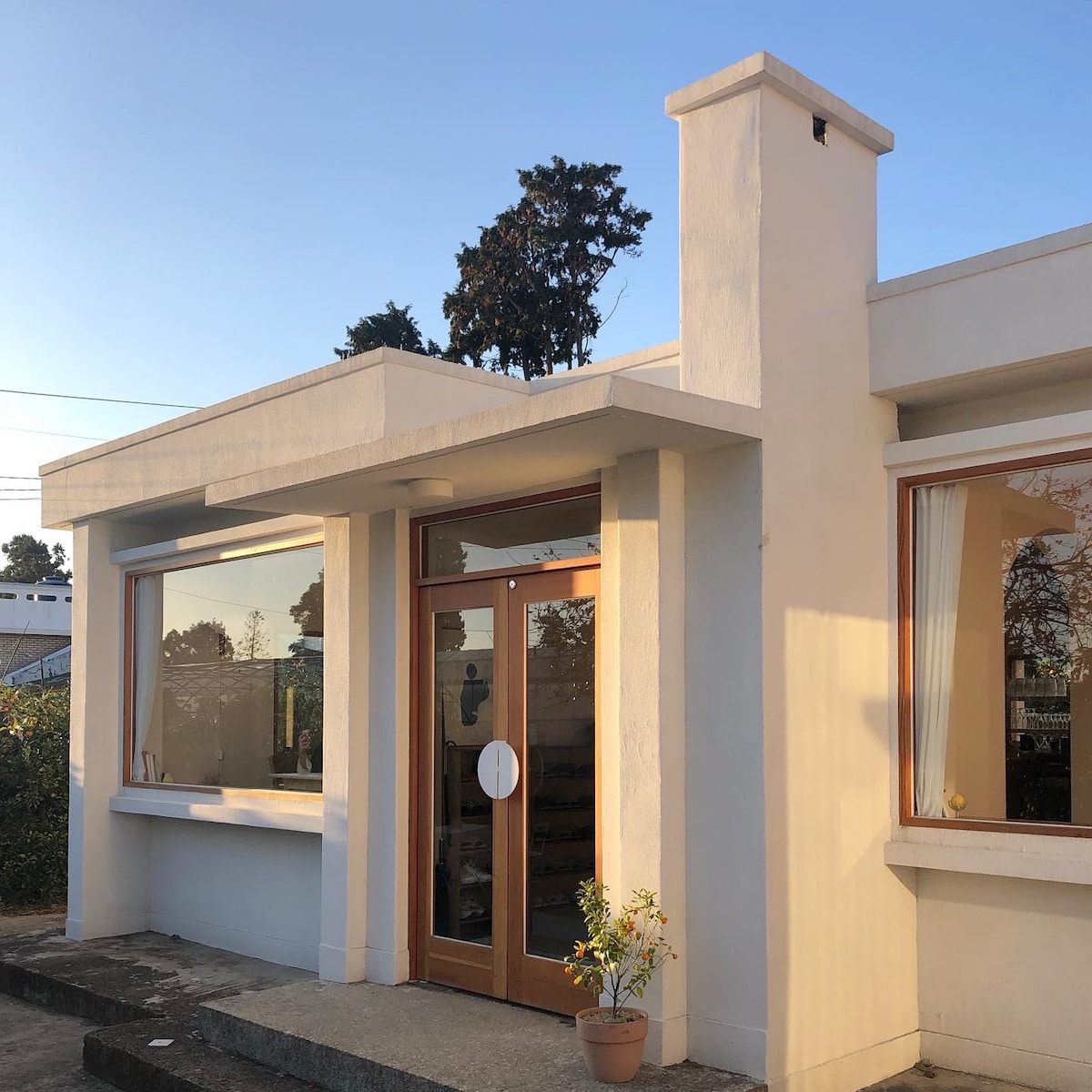 The exterior of the home is shown, featuring a clean, modern design with large windows. A wooden door with a circular handle provides an inviting entrance. A potted plant sits near the door, and trees can be seen against a clear blue sky.