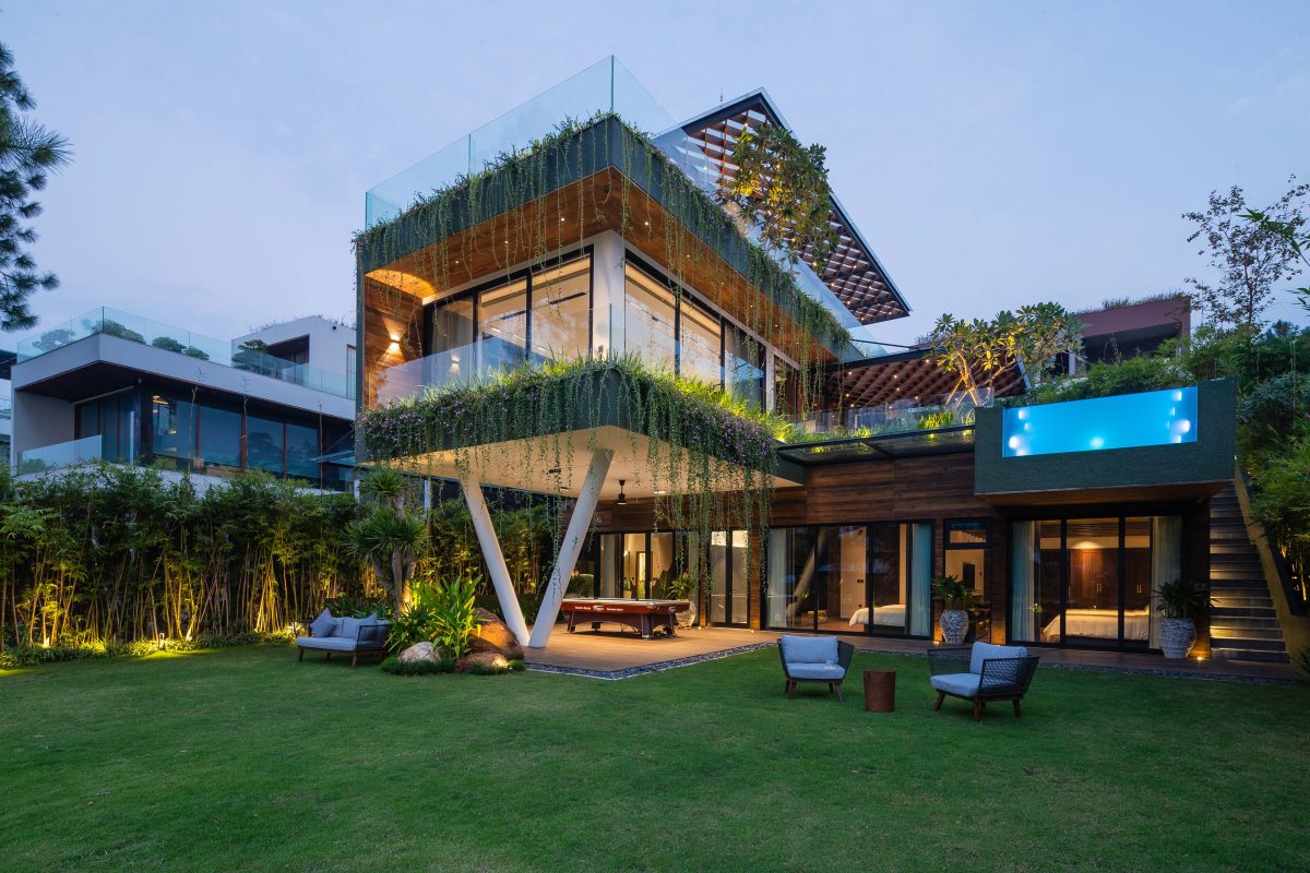 A modern house features multiple levels, with extensive glass walls and greenery integrated into the design. A spacious lawn surrounds the home, accented by outdoor seating arrangements. Soft evening light enhances the contemporary aesthetic, creating an inviting atmosphere for relaxation.