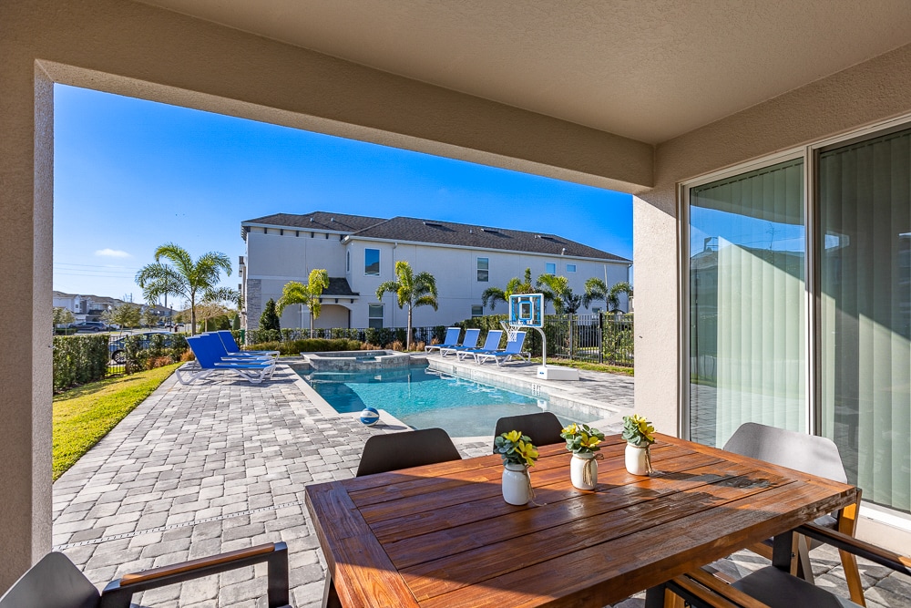 An outdoor dining table with chairs is positioned under a covered patio, overlooking a private pool with chaise lounges nearby. The pool area is surrounded by lush greenery, and a basketball hoop stands adjacent, creating an inviting space for leisure and relaxation.