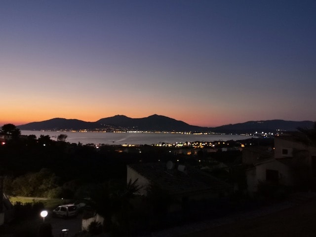 A serene twilight view is presented, featuring distant mountains with soft hues of orange and purple blending into the sky. The coastline reflects twinkling lights from nearby settlements, while a calm sea rests in the foreground.