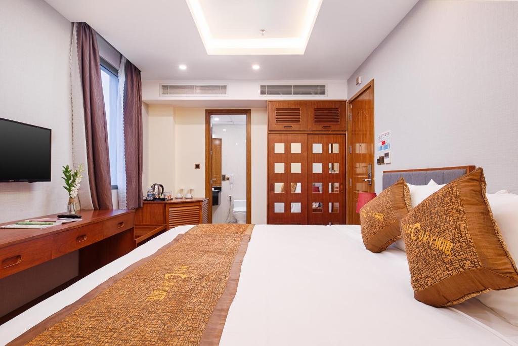 A guest room is presented with a spacious bed adorned with decorative pillows. A flat-screen TV is mounted on the wall, and a wooden desk is visible beside a kettle. Natural light enters through a window draped with curtains, illuminating the modern decor.