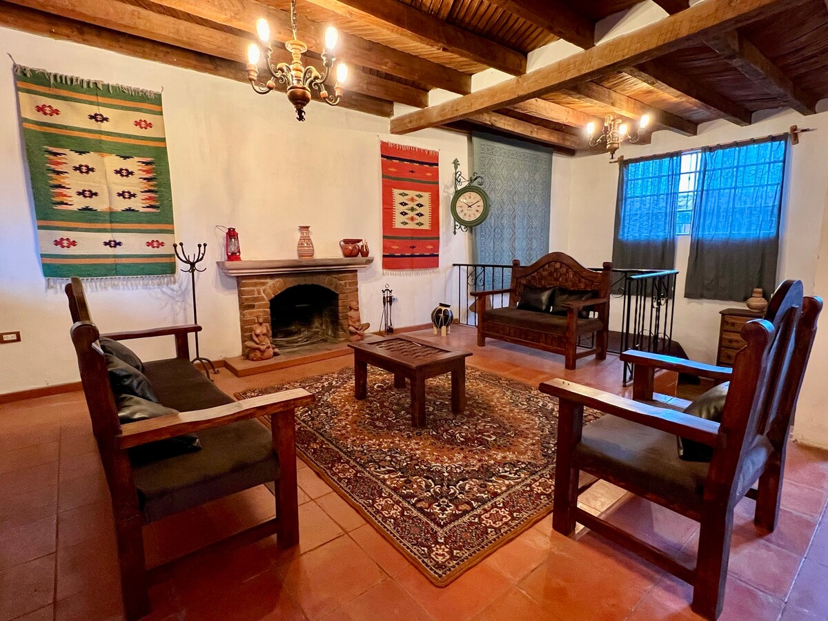A cozy living area features rustic wooden furniture arranged around a central table. Colorful textile wall hangings add character, while natural light filters through curtains. A stone fireplace serves as a focal point, enhancing the inviting ambiance of the space.