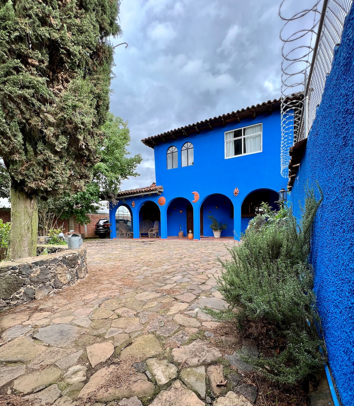 A vibrant blue house features arched entries adorned with decorative elements. The stone-paved area is surrounded by greenery, creating a serene outdoor space. A large tree and low shrubs enhance the property's natural feel, with cloudy skies visible overhead.
