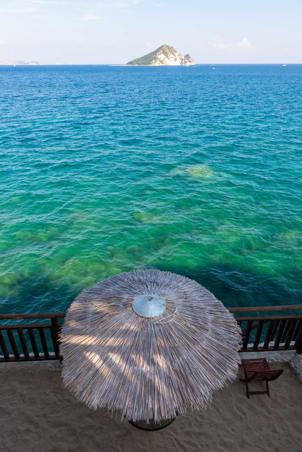 A thatched umbrella provides shade on a sandy area, overlooking clear turquoise waters. An island is visible in the distance, surrounded by the sea. A wooden railing frames the scene, adding a touch of structure against the natural backdrop.
