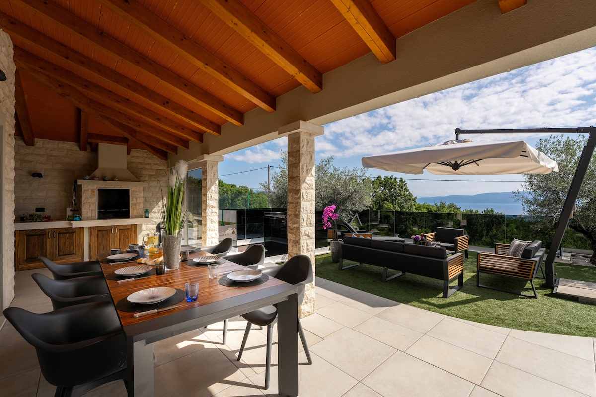 An outdoor dining area features a wooden table set for eight, accompanied by stylish black chairs. Nearby, a lounge area with a sectional sofa is positioned under a large umbrella, offering scenic views of greenery and the distant sea through a glass railing.