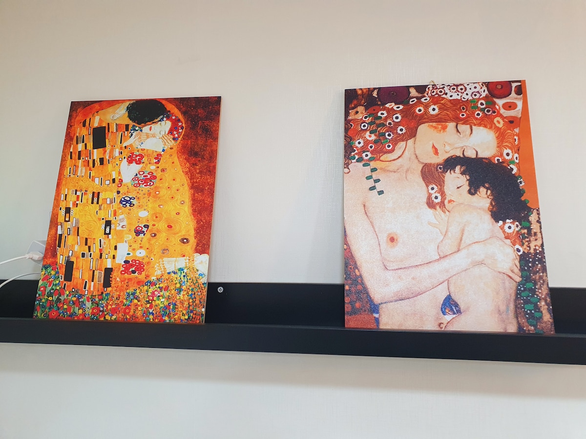 Two art pieces are displayed on a wall-mounted shelf. The left piece features abstract patterns with warm colors and floral motifs, while the right piece depicts a tender embrace between a mother and child, highlighted with soft colors and intricate details.