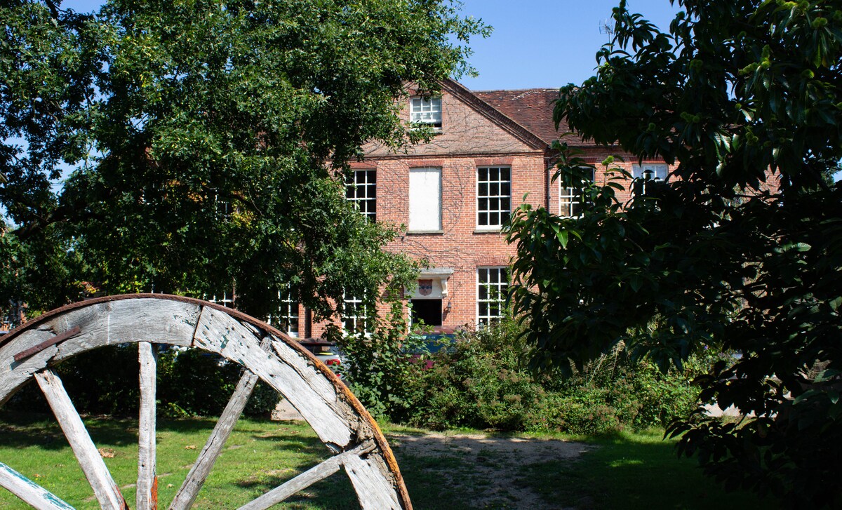 The exterior of a red brick building is framed by lush greenery and trees, creating a serene atmosphere. A vintage wooden wheel is positioned in the foreground, adding character to the scene. Large windows allow natural light to fill the interior.