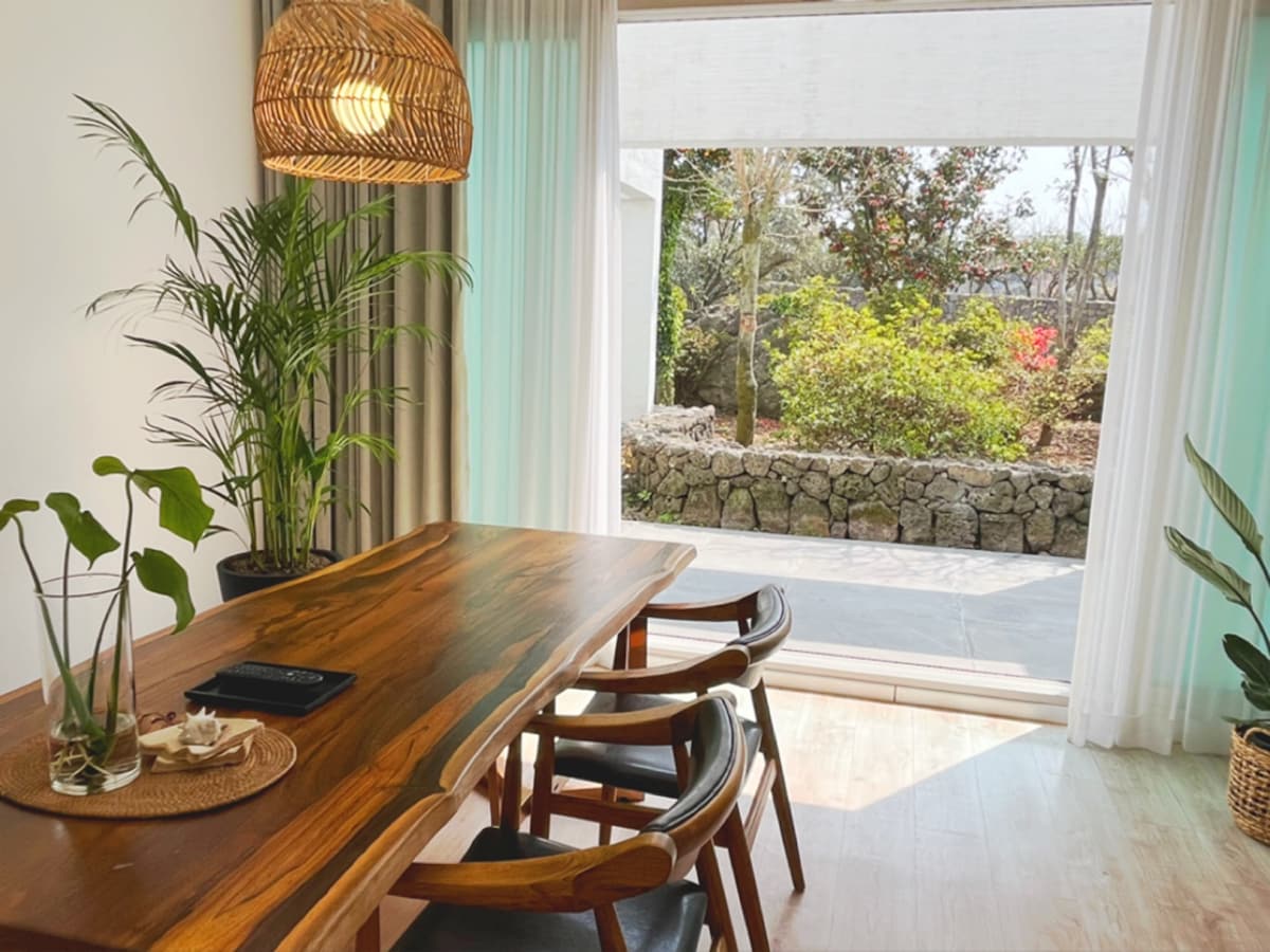 A dining area is adorned with a natural wood table, complemented by dark wooden chairs. A woven pendant light hangs above, and large glass doors reveal a lush garden view. Tropical plants are placed near the windows, adding a refreshing touch to the space.