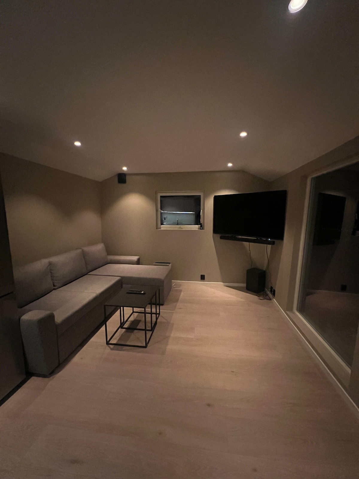 A cozy living area features a gray sofa facing a wall-mounted television. A small black coffee table occupies the center, positioned on light wooden flooring. Ambient lighting fixtures are placed in the ceiling, and a window allows for natural light to enter the space.