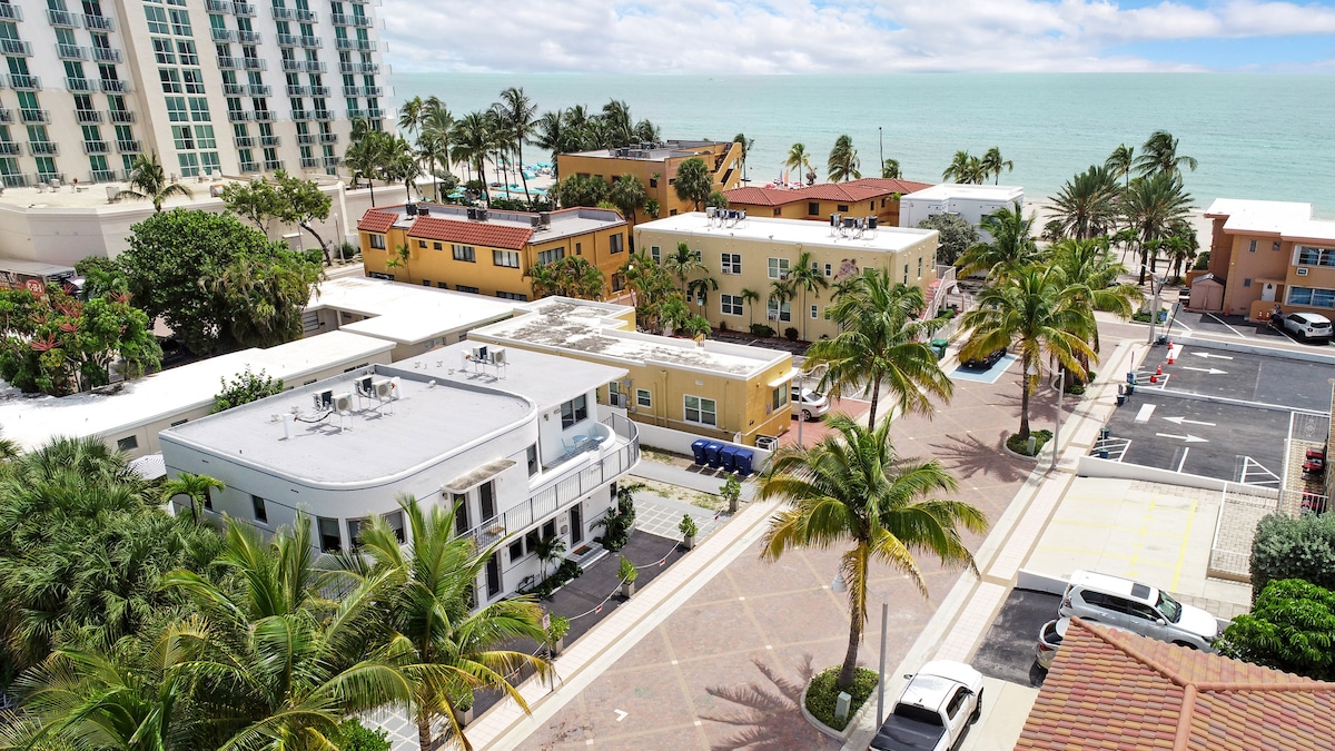An aerial view captures a vibrant neighborhood lined with palm trees, showcasing colorful buildings and a glimpse of the ocean in the background. The layout reveals designated parking areas and a well-maintained pathway, highlighting the proximity to the beach and surrounding establishments.