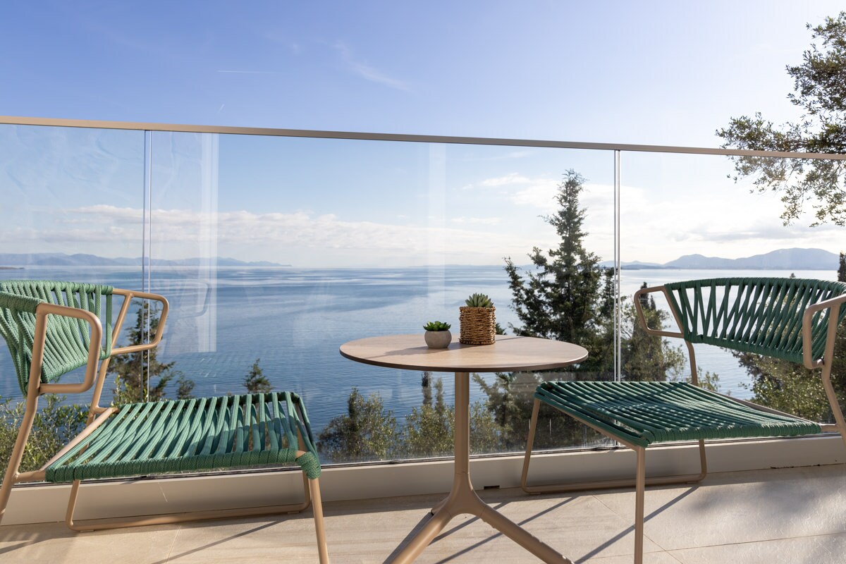 A balcony area is presented with two green chairs and a round table, enhancing the outdoor space. The glass railing offers an unobstructed view of the sea and the surrounding landscape, allowing for an inviting atmosphere to enjoy the serene surroundings.