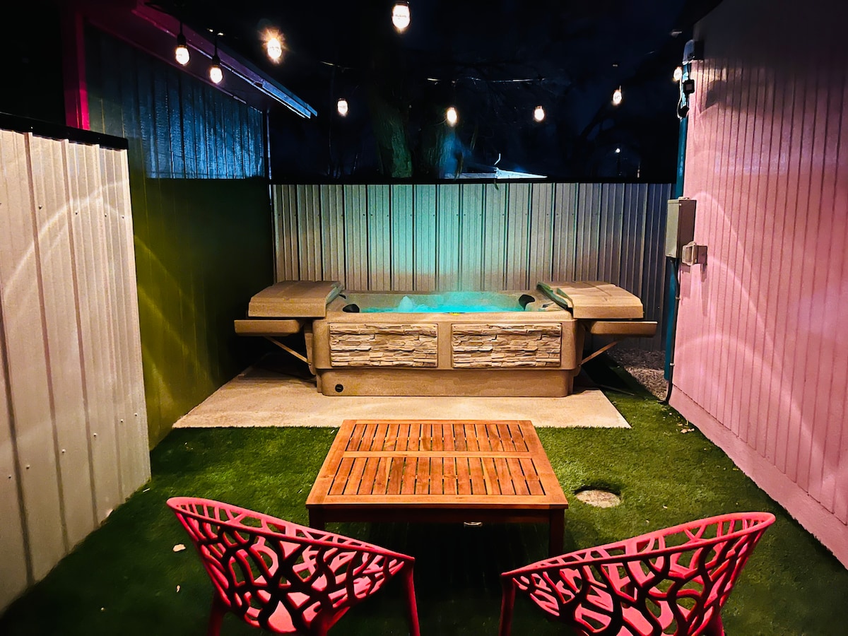 A private outdoor space features a hot tub with stone accents under softly glowing string lights. Two vibrant pink chairs are positioned around a wooden coffee table on the green grass, creating a space ideal for relaxation and socializing.