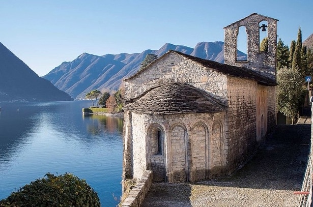 An ancient stone church stands beside the serene waters of Lake Como, its rounded windows and bell tower visible against a backdrop of mountains. The calm lake reflects the surrounding landscape, while a pathway leads to the church, bordered by trees and natural foliage.