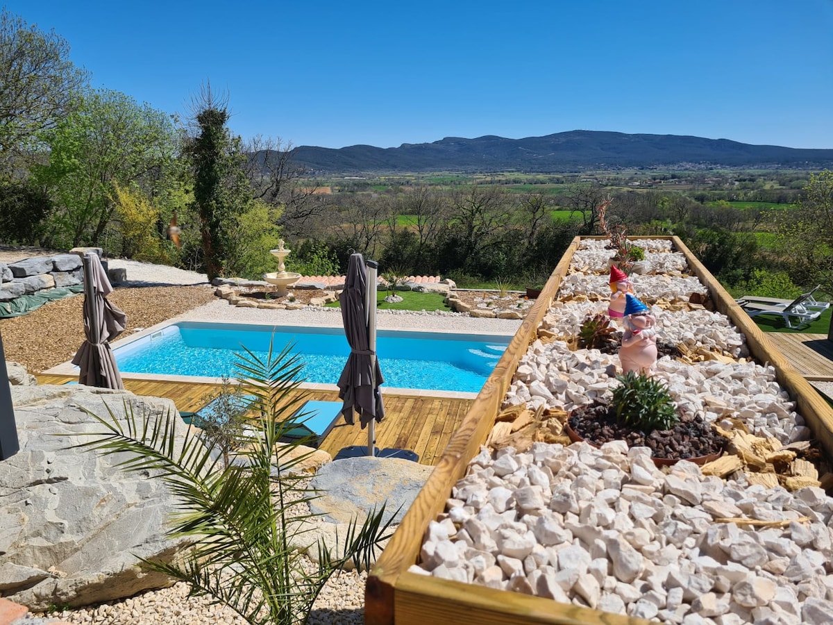 A serene outdoor area features a heated swimming pool surrounded by landscaped gardening stones and plants. Comfortable lounge chairs are positioned near the pool, with panoramic views of rolling hills and countryside visible in the background under a clear blue sky.