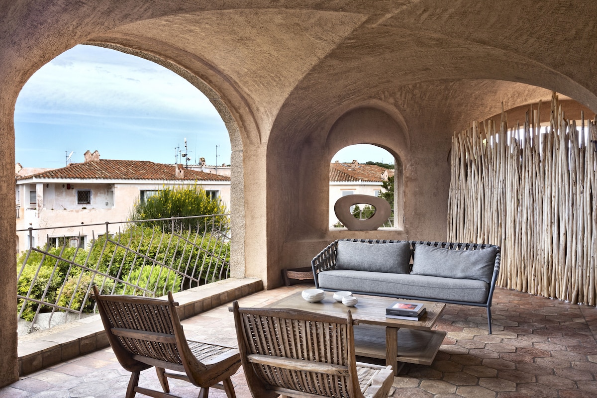 An arched balcony area features a grey sofa accompanied by two wooden armchairs, offering a comfortable sitting arrangement. A coffee table is positioned in front, with natural light enhancing the neutral tones. The backdrop includes residential buildings and greenery, framed by the architectural arches.