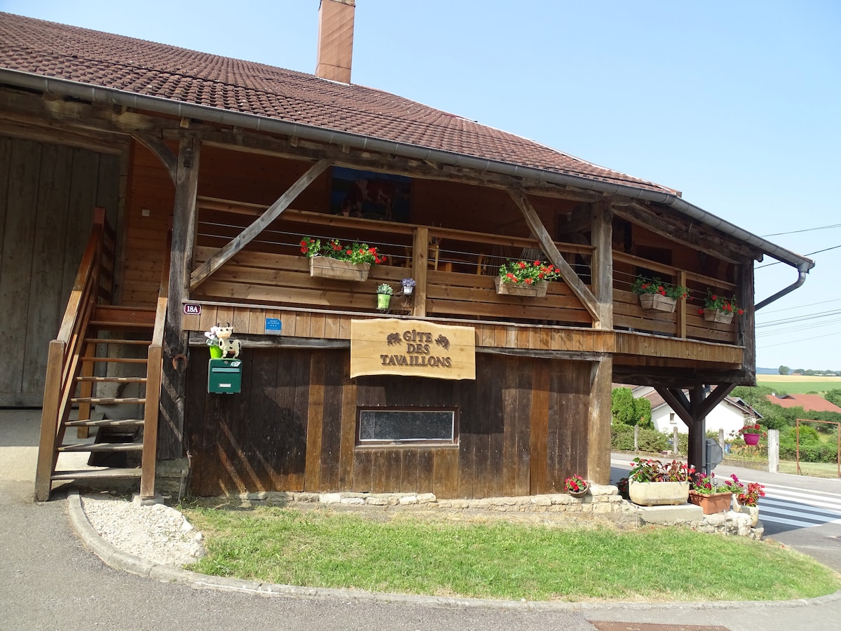 The exterior of the gîte features rustic wooden architecture and a sloped roof. Flower boxes filled with colorful blooms adorn the balcony, while a wooden sign identifies the property. Stairs lead to the entrance, and the surroundings include a paved path and landscaped areas.
