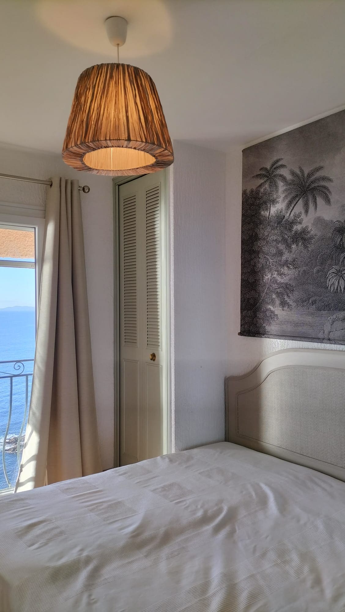 A bedroom features a light-colored headboard and a simple bedside light fixture with a woven shade. Soft light enters through a window adorned with draped curtains, while a mural depicting tropical foliage enhances the room's ambiance.