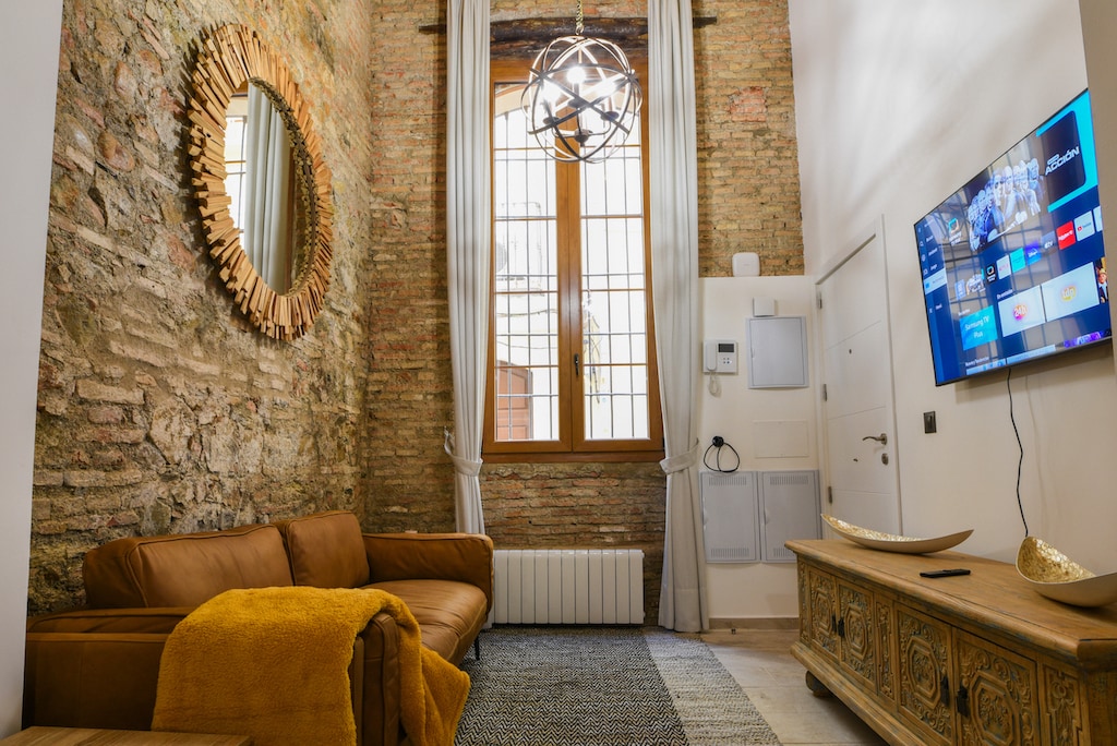 A living area features exposed brick walls that add character, complemented by a brown leather sofa with a bright yellow throw blanket. A round mirror reflects natural light from the large window, while a modern light fixture hangs above. A television is mounted on the wall opposite.