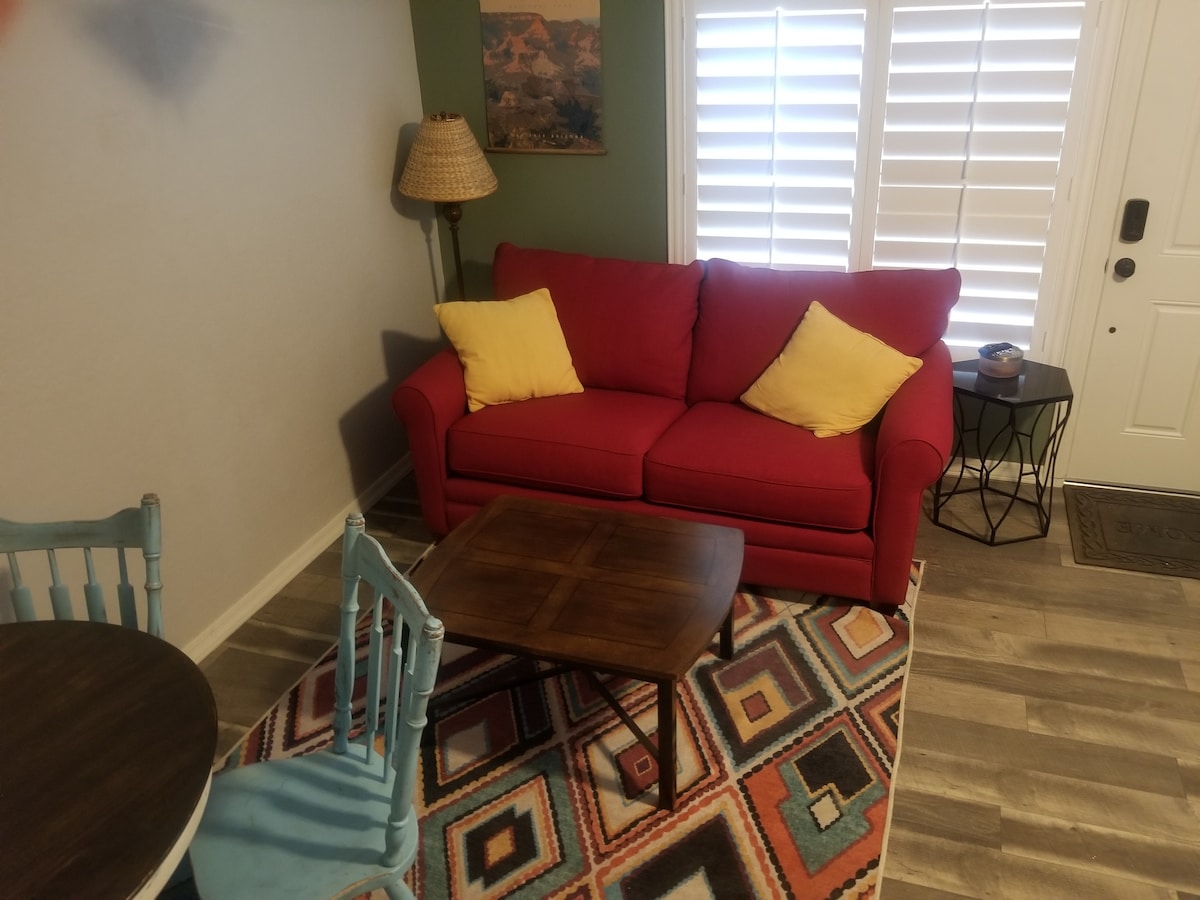 A welcoming living area displays a vibrant red sofa adorned with yellow pillows. A rustic coffee table sits on a patterned rug, complemented by a small dining table with blue chairs. Light filters through plantation shutters on the windows, enhancing the neutral color scheme.