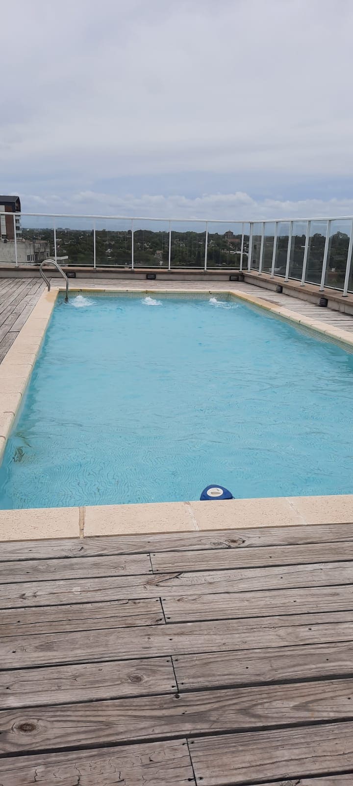 An outdoor swimming pool is displayed, surrounded by a wooden deck. The clear blue water reflects the sky, and a safety buoy floats near the edge. A glass railing provides an unobstructed view of the landscape beyond.
