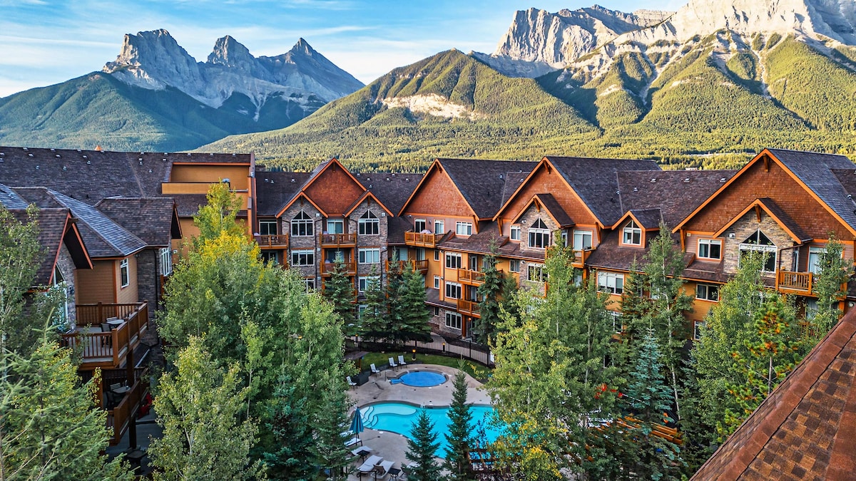 A scenic view captures the resort set amidst lush greenery, with the outdoor heated pool visible at the center. The backdrop features the majestic Canadian Rockies, while the rustic architecture of the buildings complements the natural landscape.