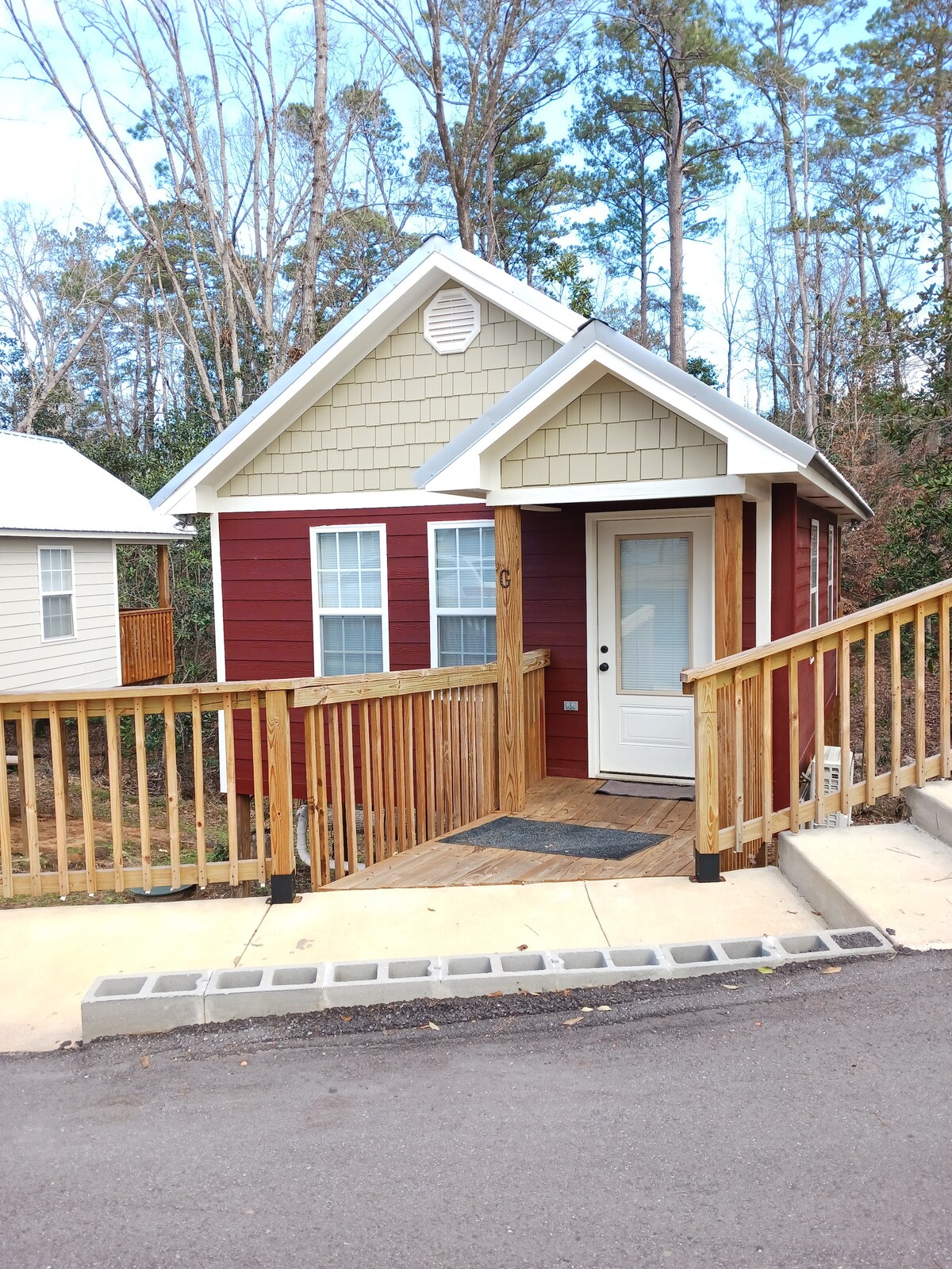 The exterior of a cozy cottage is shown, featuring a red and tan wooden facade. A welcoming front door, flanked by angular rooflines, is accessible via a wooden ramp. Surrounding trees provide a natural backdrop, enhancing the cottage's secluded feel.