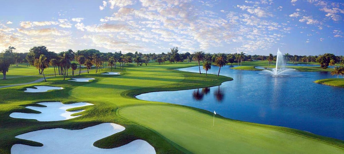 A scenic view of a golf course is presented, featuring lush green fairways, strategic sand traps, and a serene water hazard with a fountain. Palm trees are visible in the background, complemented by a clear blue sky and scattered clouds.