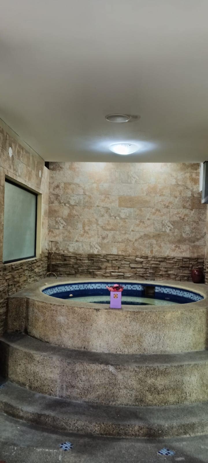 A cozy space features a circular jacuzzi surrounded by textured stone walls. Soft lighting illuminates the area, highlighting the smooth surfaces and inviting ambiance. A small decorative item, with a visible label, sits at the edge of the jacuzzi, adding a touch of detail to the setting.