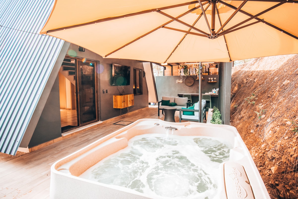 An outdoor jacuzzi is set under a large umbrella, providing a shaded area for relaxation. Next to it, a seating arrangement with modern furniture invites guests to enjoy the serene surroundings. The building's architectural design includes angled walls that complement the natural landscape.