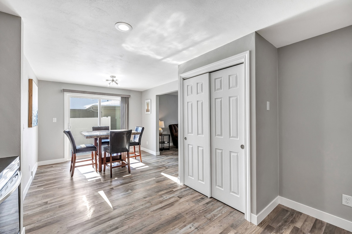 An open living space features natural light filtering through large windows. A dining table with six chairs is positioned beside a spacious closet. Light wood flooring adds warmth to the neutral-toned walls, creating a versatile area for relaxation and dining.