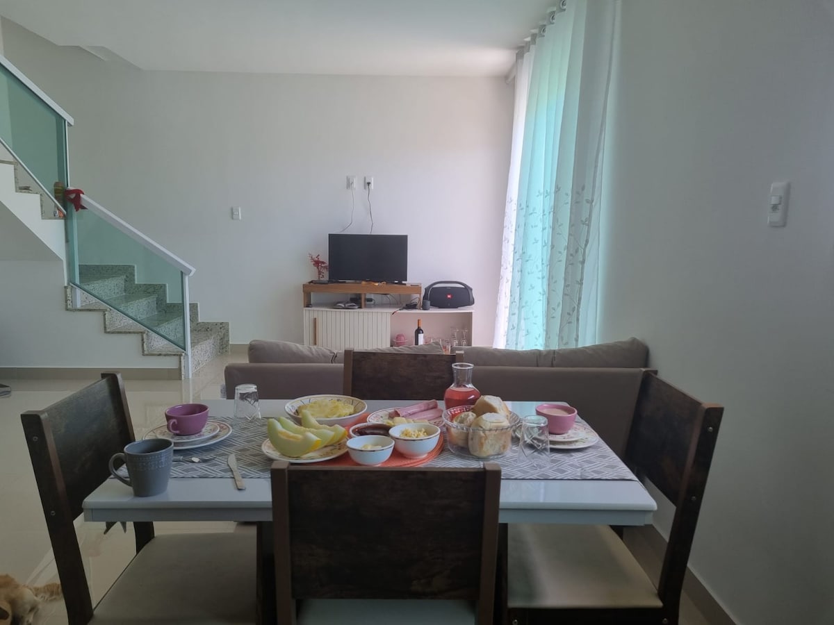 A dining table is set with various breakfast items, including plates, cups, and a glass bottle. In the background, a sofa is visible along with a television, while large windows allow natural light to brighten the space.