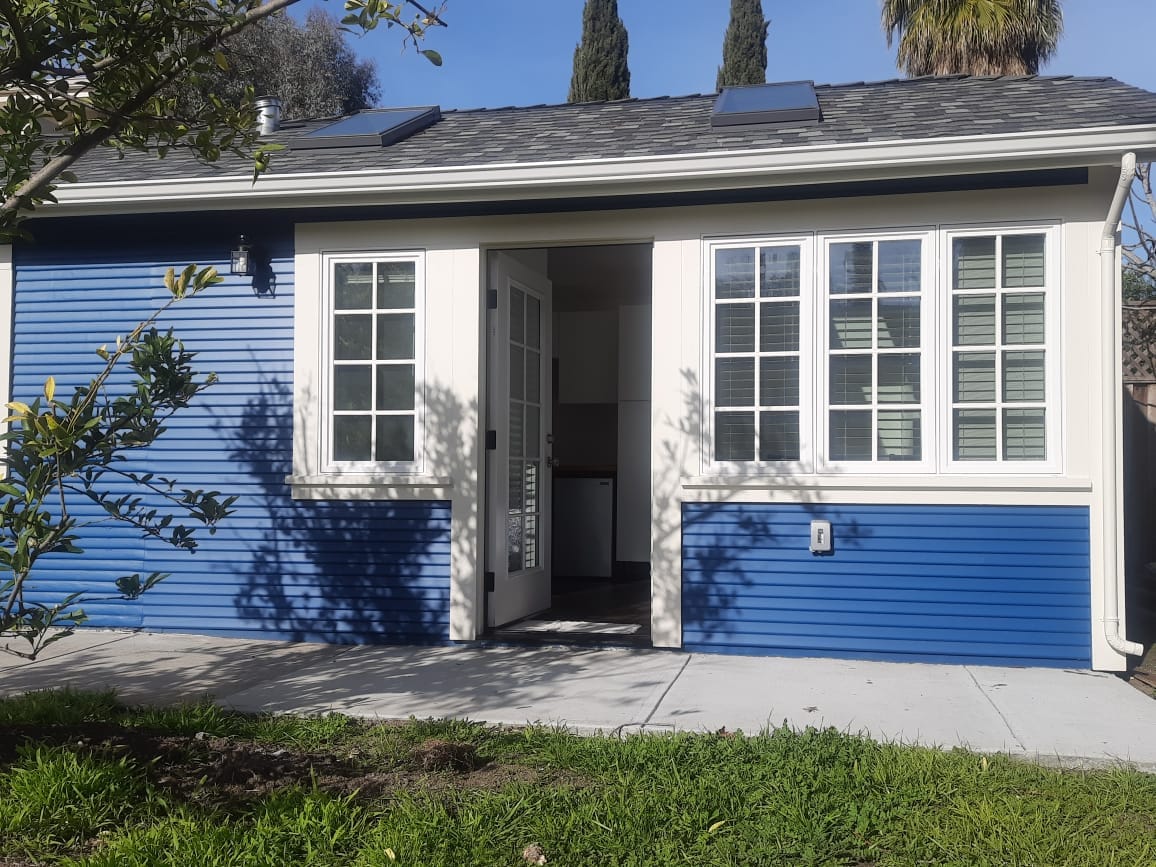 A charming blue exterior of a guest studio is showcased, featuring large windows that allow natural light. French doors provide access to the patio, with a well-maintained path leading to a grassy area adorned with greenery.