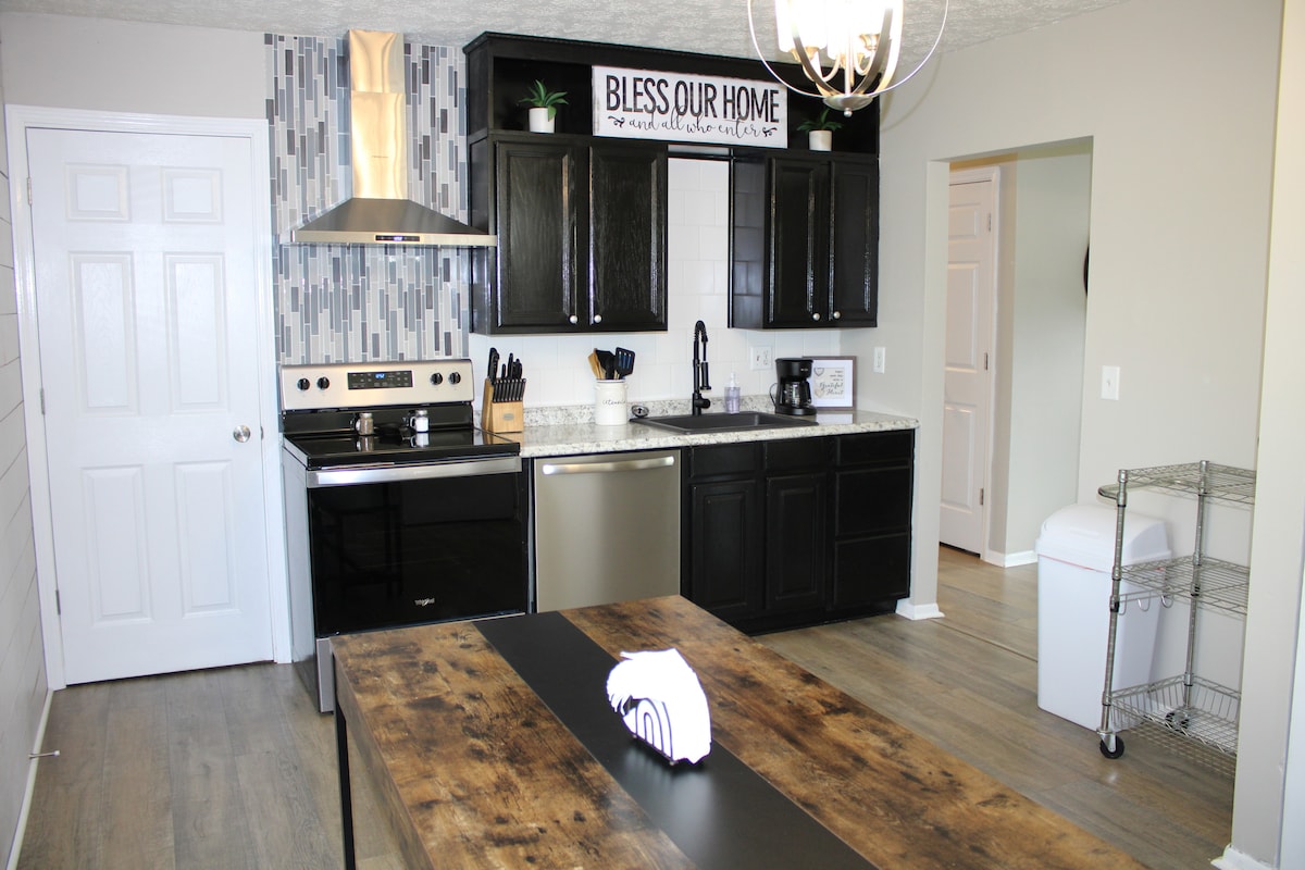 A spacious kitchen features black cabinetry and stainless steel appliances, including a stove and dishwasher. A central wooden island provides ample workspace. Decorative elements, such as a sign reading 'Bless Our Home,' add character to the space, complemented by natural light from a nearby entrance.