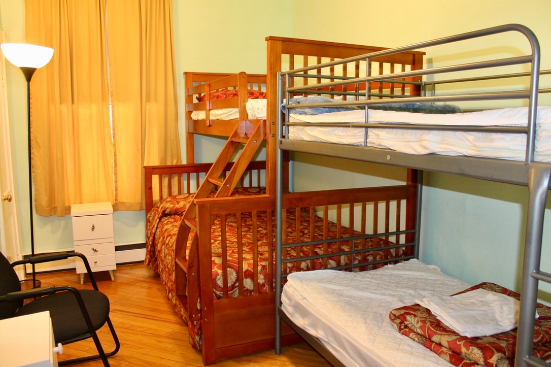 The room features a set of four bunk beds, including one double bed on the bottom and three single beds above. Soft, patterned bedding and light-colored walls contribute to the space. A window is adorned with light curtains, allowing natural light to illuminate the area.