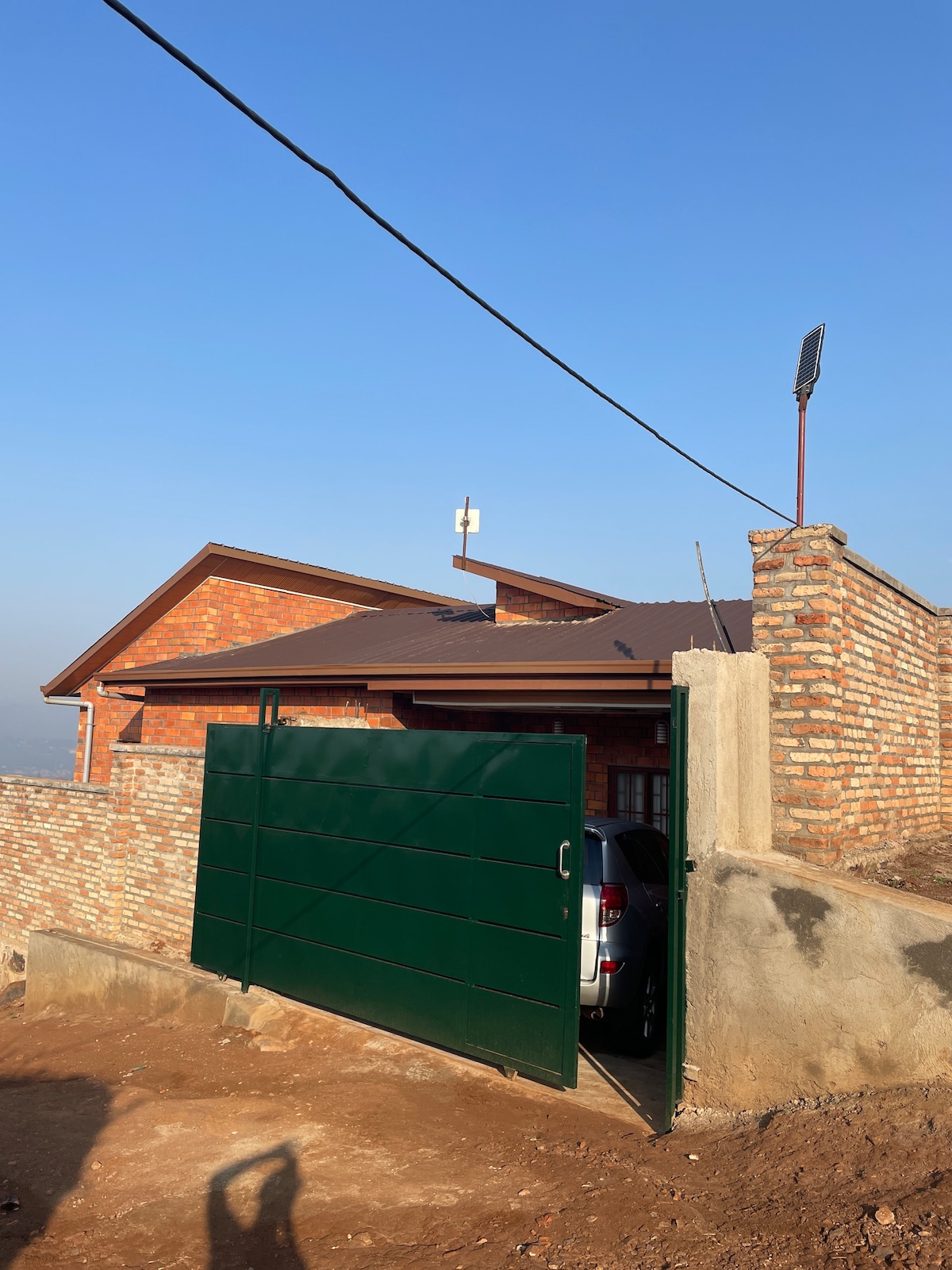 A green gate opens into a private parking area, showcasing a vehicle and the sturdy brick structure of the home in the background. The clear blue sky above enhances the peaceful setting.