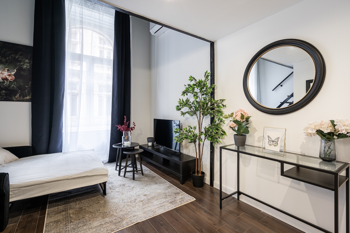 A modern living space is highlighted by a minimalist design, featuring a large mirror and a sleek flat-screen television mounted on a dark furniture unit. A small dining table is placed next to a decorative plant, while floral arrangements are displayed on a glass console.