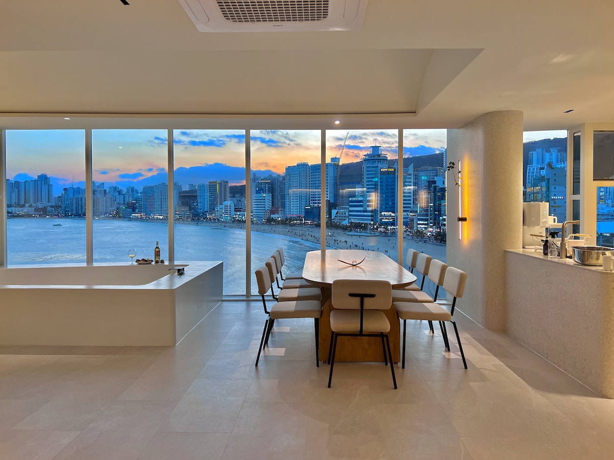 A dining area features a long table surrounded by chairs, with large windows showcasing an ocean view and city skyline. The soft glow of the setting sun casts a warm light across the space, creating a serene dining atmosphere.