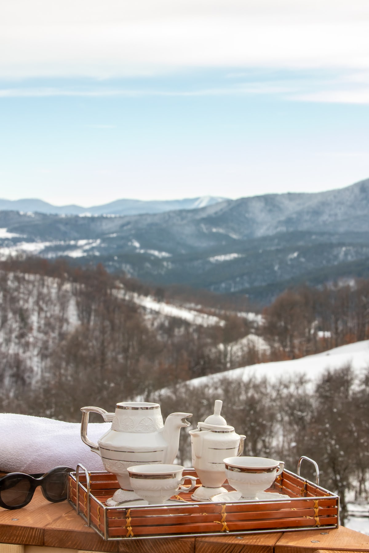 A wooden tray is positioned on a table, holding a white tea set that includes a teapot, sugar bowl, and cup. Snow-covered mountains and clear skies serve as a serene backdrop, creating a peaceful winter scene.