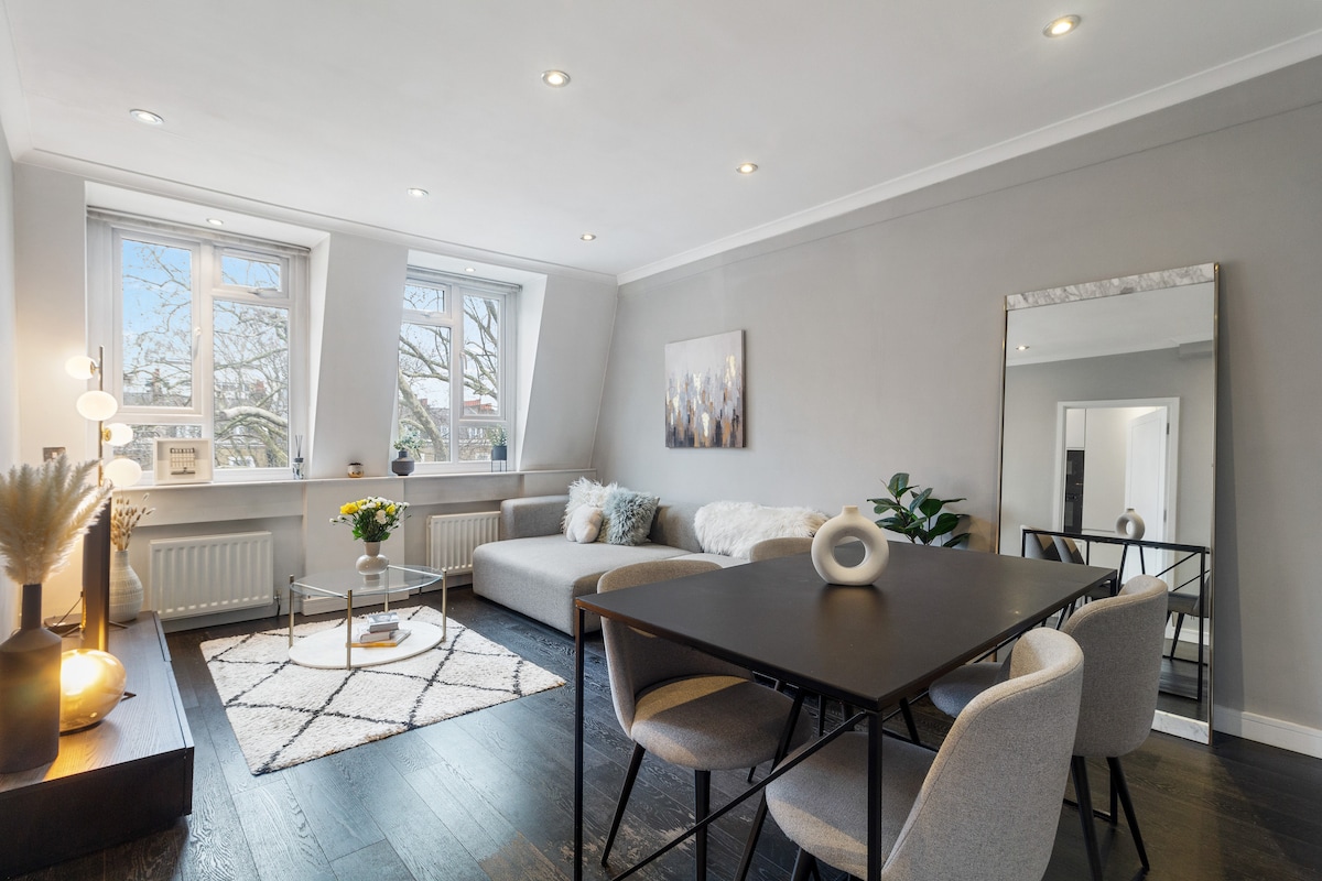 A modern living area is featured with a spacious sofa and large windows allowing natural light to fill the space. A contemporary dining table with four chairs sits beside a stylish rug, complemented by a decorative mirror and plants, creating a welcoming ambiance.