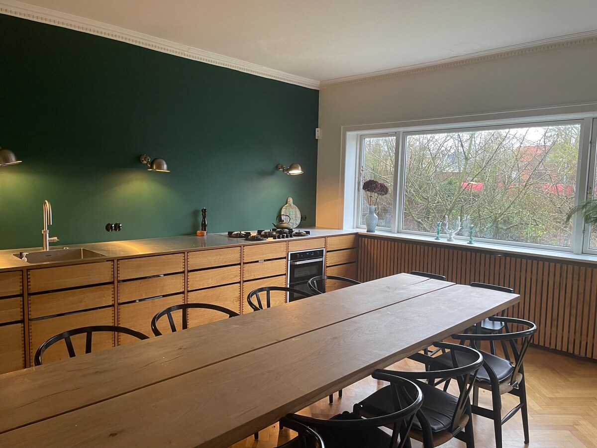 The kitchen features a long wooden table surrounded by black chairs. A green accent wall is complemented by warm wooden cabinetry and a large window that offers natural light and views of the outside greenery. Modern appliances are integrated into the design.