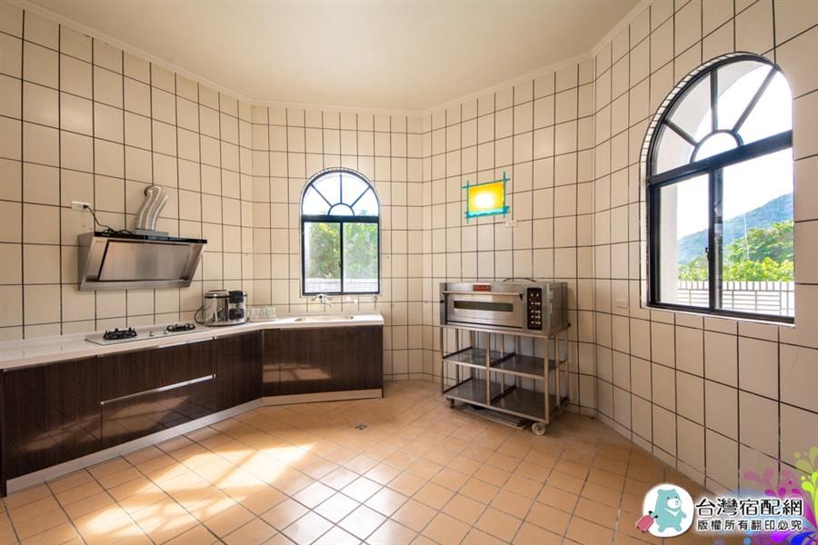 A spacious kitchen is presented with tiled walls and a tiled floor. Large windows allow natural light to fill the area. A modern cooking area features an extractor hood above a stovetop, while a metal cart stands adjacent to an oven, providing ample space for food preparation.