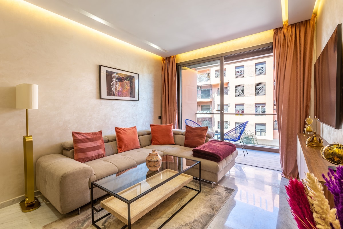 A modern living room features a spacious sectional sofa adorned with orange and burgundy cushions. A coffee table sits atop a light-colored rug, while large glass doors open onto a balcony. Natural light frames the room, complemented by soft lighting along the ceiling.