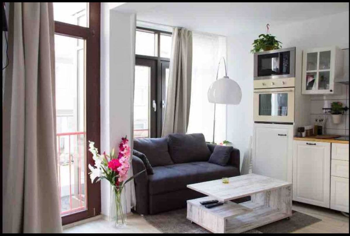 A cozy living area is presented, featuring a comfortable dark sofa and a wooden coffee table. Natural light enters through large windows adorned with elegant curtains. Potted plants add a touch of greenery, while a modern kitchen area is partially visible in the background.