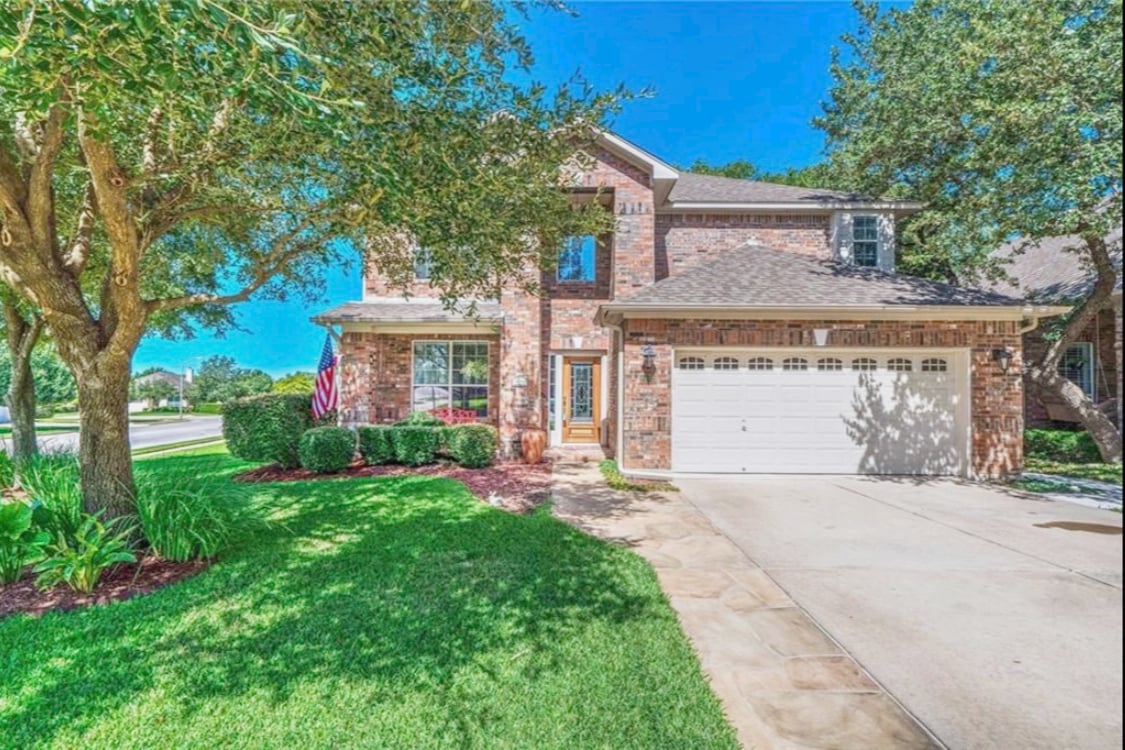 A two-story brick home is framed by lush green landscaping. A well-maintained driveway leads to a two-car garage. An American flag is displayed on the porch, welcoming guests. Sunlight filters through the trees, casting gentle shadows on the manicured lawn.