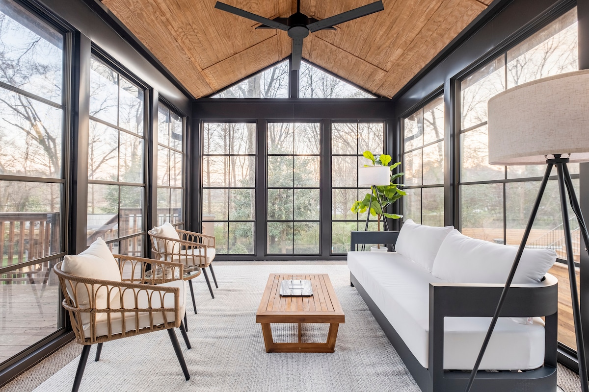 A sunroom is surrounded by large windows, allowing natural light to illuminate the space. Two chairs with a woven design are positioned beside a wooden coffee table. A white sofa adds comfort, while a standing lamp and indoor plant provide a touch of greenery.