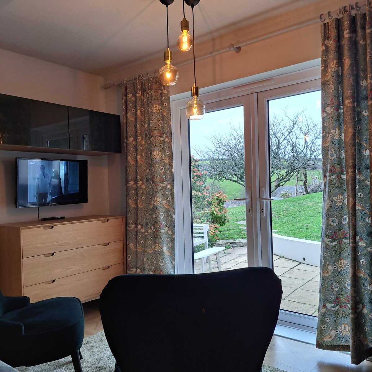 A cozy sitting area features a dark armchair and a light wooden dresser with a television on top. Large glass doors open to a patio area, while patterned curtains frame the windows. Soft lighting is provided by hanging bulbs, enhancing the inviting space.